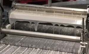 An industrial icing leveler drips icing over a conveyor belt, and an air knife blows away the excess product.